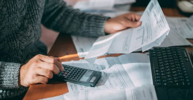 Close de mãos masculinas com uma conta de serviços públicos, vários cheques e uma calculadora sobre a mesa.
