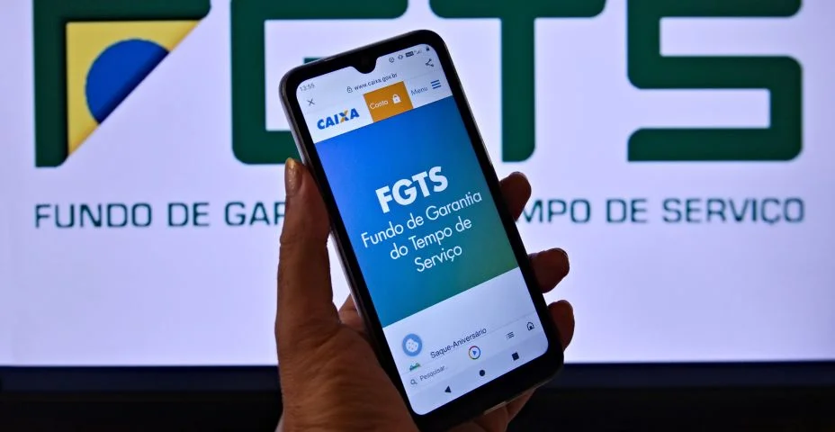 O logotipo do FGTS na tela de um celular sendo segurado por uma mão. Ao fundo,