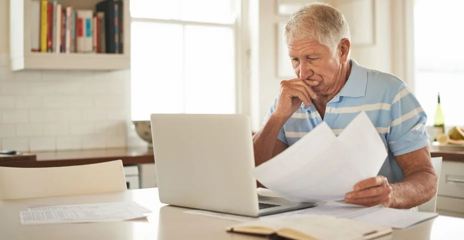 senhor olhando para o notebook enquanto está segurando varios papeis