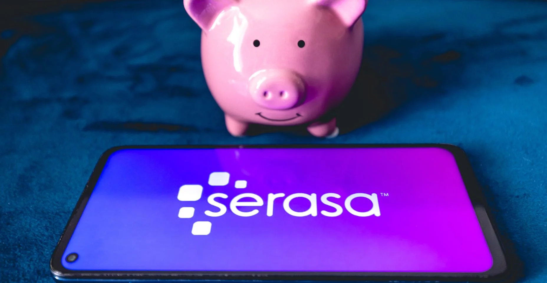 Um telefone celular com a logo da empresa brasileira Serasa e um cofrinho sobre um sofá azul. Consulta, inadimplência, consumidor, dívida vencida, nome sujo, negativado, limpar o nome.