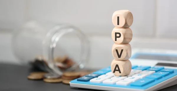 A sigla IPVA escrita em dados de madeira que estão sobre uma calculadora. Um pote de vidro com moedas do Brasil na composição.