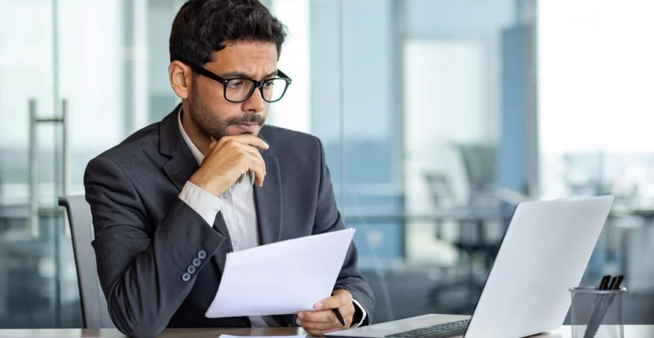 Retrato de empresário sério pensativo atrás de papelada, financista olhando documentos, papéis e contratos, pensando em soluções para definir tarefas, homem dentro do escritório em terno de negócios com laptop