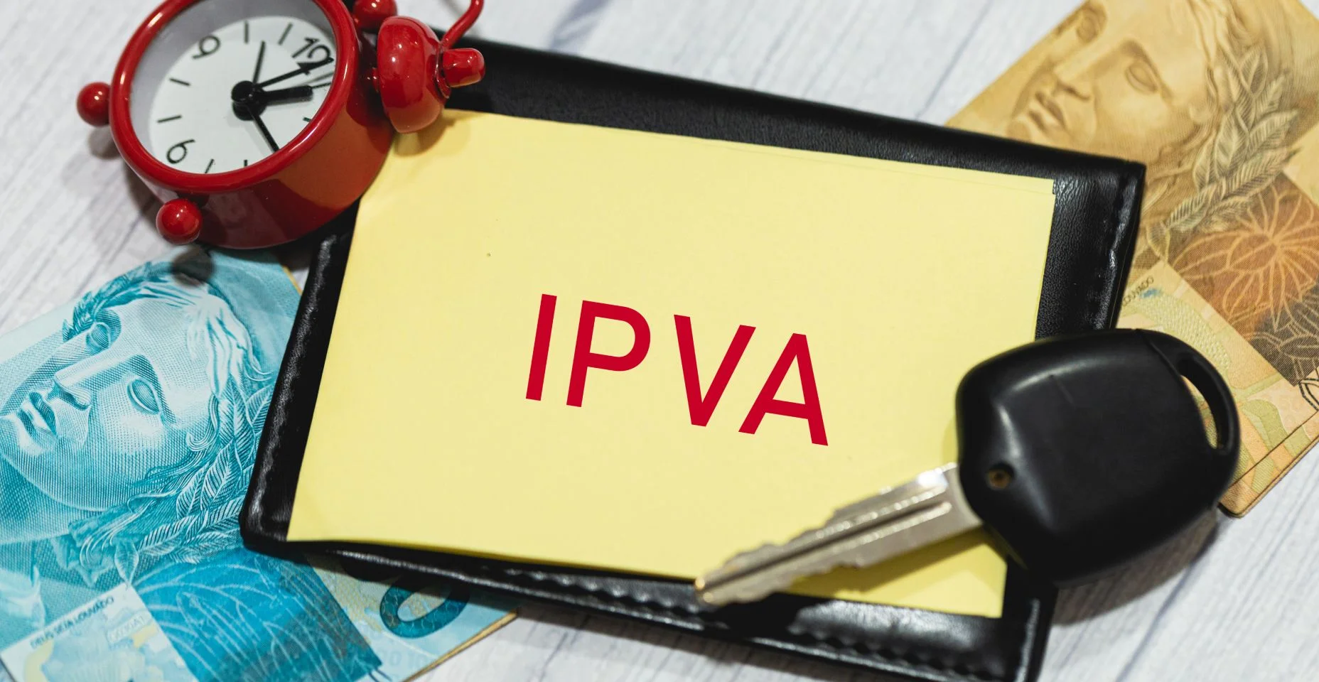 A sigla IPVA refere-se ao Imposto sobre a Propriedade de Veículos Automotores em português do Brasil escrito em um pedaço de papel. Notas de reais, chave do carro e portador de documento na composição.