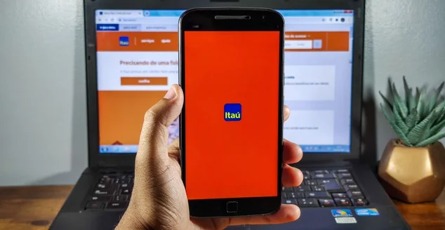 homem segurando smartphone com aplicativo do Banco Itaú na tela, com computador ao fundo. Banco brasileiro.