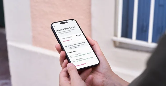 Uma pessoa segura um smartphone, cuja tela exibe uma interface de aplicativo em português. O aplicativo mostra informações do "Serasa e Vale Bonus", incluindo "Saldo acumulado: R$ 7,50", "Limite de bônus do Serasa: R$50", e uma lista indicando que o usuário ganhou três vales-presente. Há também botões como "Utilizar saldo >" e "Ver vales completos >".