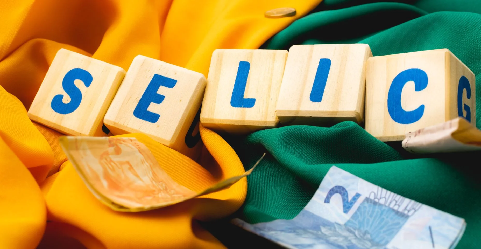 Palavra SELIC escrita em cubos de madeira com algumas cédulas do Real Brasileiro. Fundo em cores amarelo e verde associando ao Brasil.