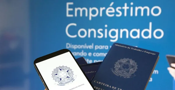 Imagem com fundo azul e o texto 'Empréstimo Consignado' em destaque. Abaixo, há a frase 'Disponível para ...'. Em primeiro plano, aparecem duas Carteiras de Trabalho e Previdência Social — uma física e outra exibida na tela de um smartphone.