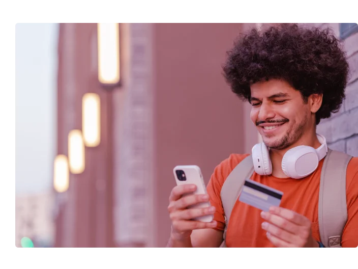 Estudante jovem olhando para o celular sorrindo, com o cartão de crédito na outra mão