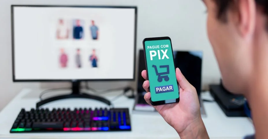 Mãos segurando smartphone para pagar compras online via Pix. Compras online no computador e uso de aplicativo móvel para pagamento instantâneo
