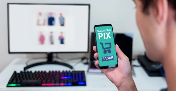 Mãos segurando smartphone para pagar compras online via Pix. Compras online no computador e uso de aplicativo móvel para pagamento instantâneo