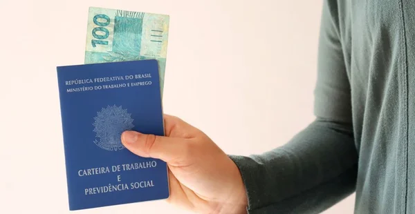 Pessoa segurando carteira de trabalho brasileira e cem reais.