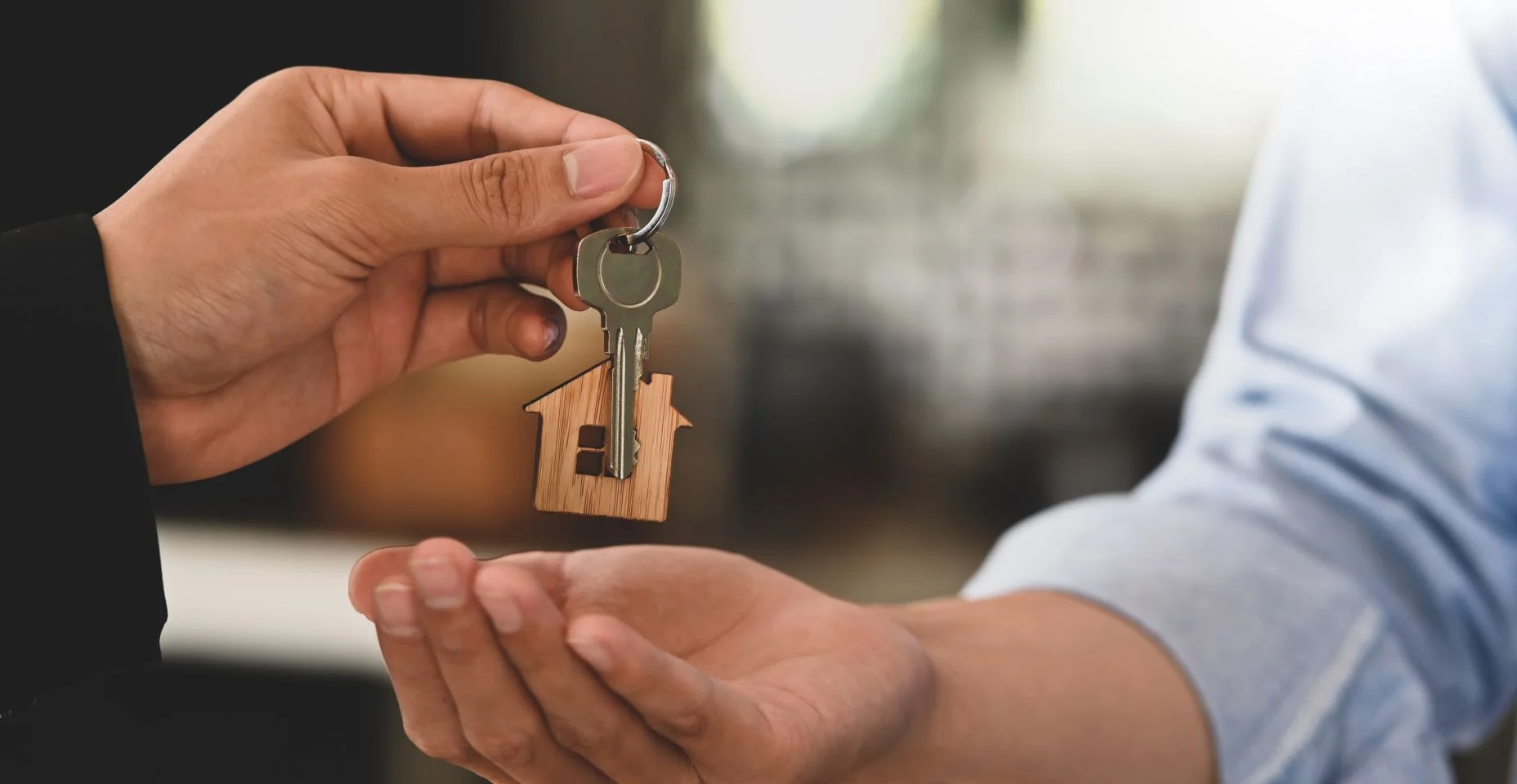 Imagem recortada Mãos de um corretor de imóveis oferecendo / dando uma chave de casa para um homem inteligente de camisa azul enquanto estão juntos sobre um banco moderno como pano de fundo. Conceito de corretor/vendedor/revendedor.