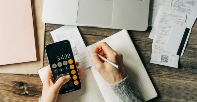 Vista superior de uma mesa de trabalho na qual uma mulher mantém um orçamento usando uma calculadora.