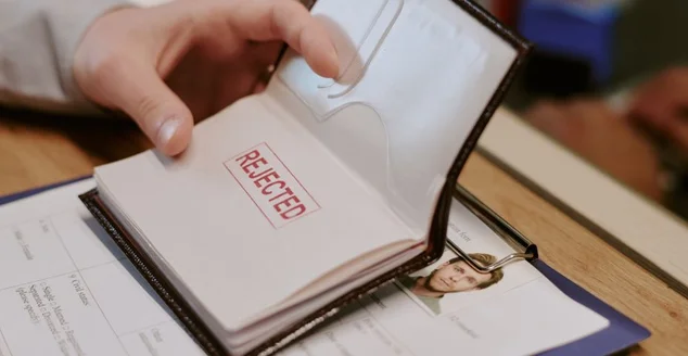 Mão abrindo passaporte exibindo grande carimbo vermelho de rejeição na página, acompanhado de alguns documentos pessoais e prancheta com papéis sobre a mesa