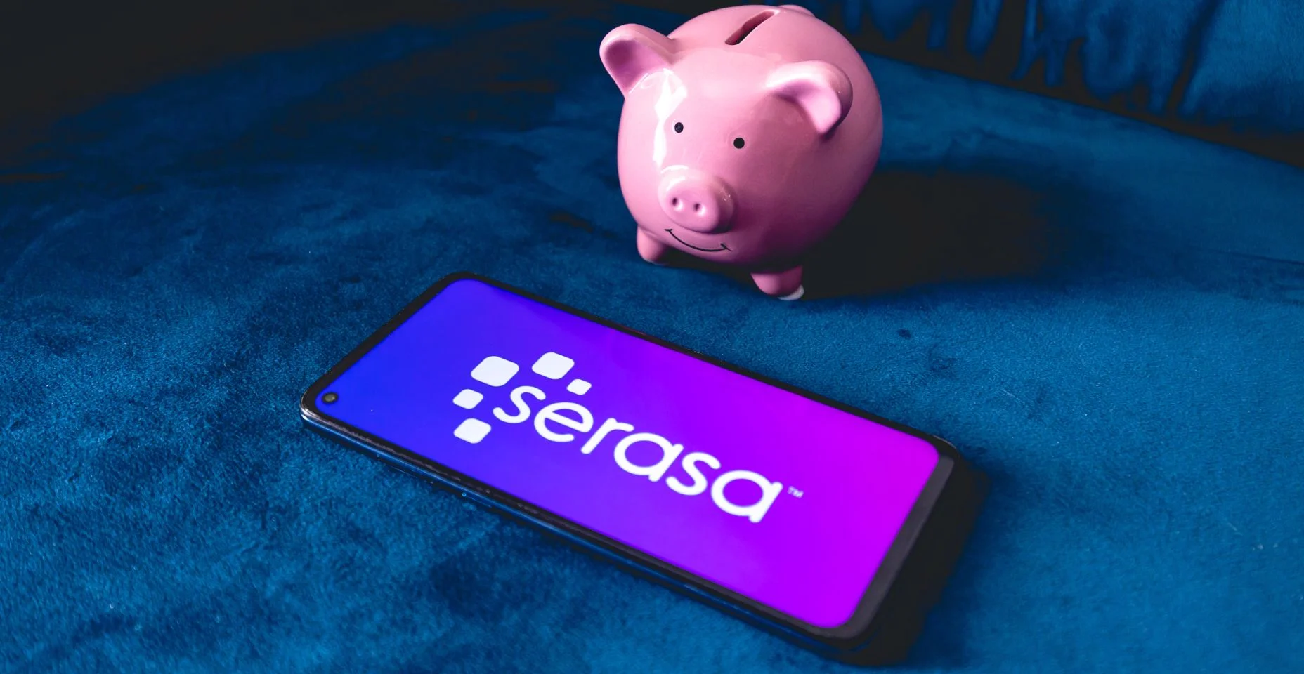 Um telefone celular com a logo da empresa brasileira Serasa e um cofrinho sobre um sofá azul.