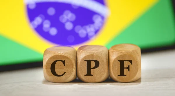 A sigla CPF para Registro de Pessoa Física em português brasileiro escrita em dados de madeira. Ao fundo, a bandeira do Brasil na tela de um celular.