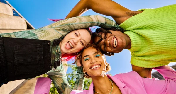 rês adultos da Geração Z formando um círculo com os braços e sorrindo ao ar livre em um ambiente urbano. Tiro de baixo ângulo.