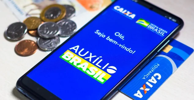 Nesta fotoilustração, o logotipo do Auxílio Brasil é exibido em um smartphone, ao lado de notas de dinheiro real brasileiro.