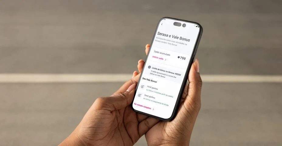 Uma pessoa segura um smartphone com as duas mãos. A tela do celular exibe um aplicativo financeiro em português, mostrando informações sobre "Serasa e Vale Bonus". São visíveis dados como "Saldo acumulado" com o valor de 750, além de opções como "Linha de bônus" e "Seus pontos".