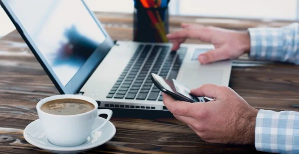 Homem mexendo no notebook com um celular na outra mão e uma xicará de café ao lado.
