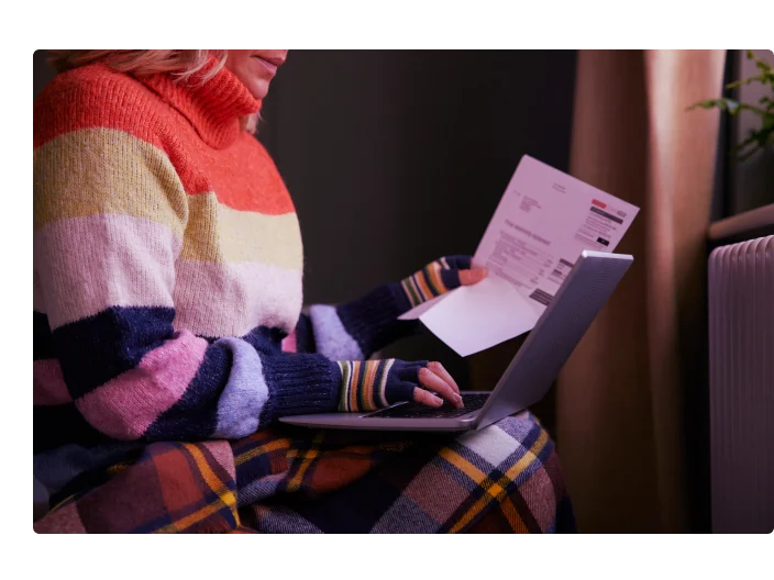 Mulher de luvas com laptop e conta tentando se manter aquecida pelo radiador durante a crise de energia do custo de vida