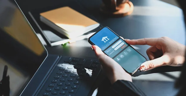 Foto recortada de uma mulher executiva segurando o smartphone nas mãos enquanto faz transações financeiras on-line por seu smartphone na frente do tablet do computador na mesa de trabalho moderna.