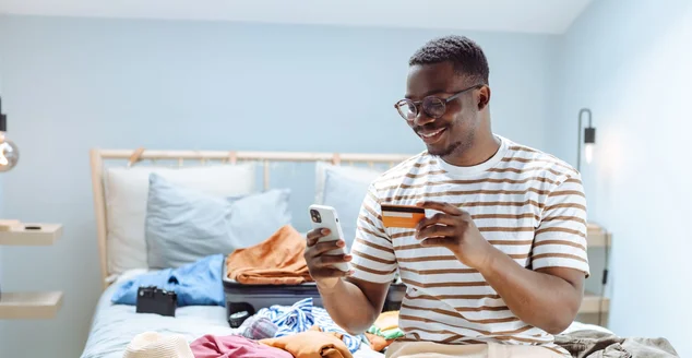 Homem afro-americano fazendo as malas para uma viagem e usando o telefone inteligente para reserva on-line