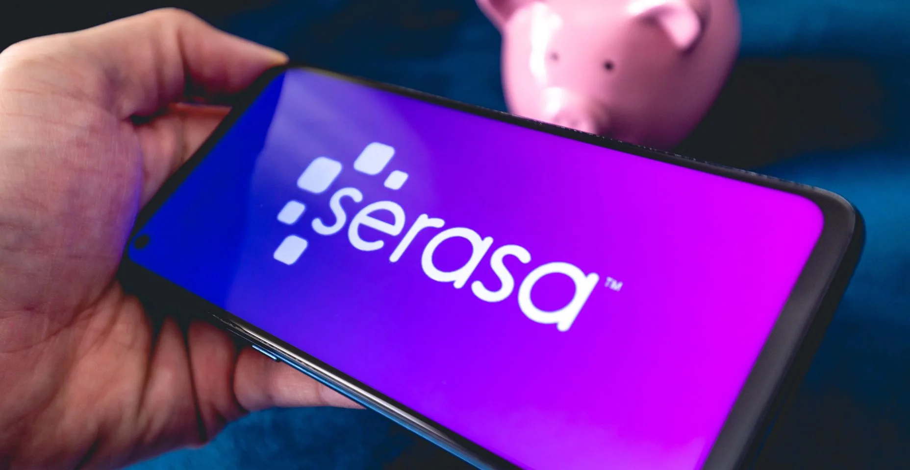 Um homem segurando um telefone celular com a logo da empresa brasileira Serasa. Um cofrinho na composição. Consulta, inadimplência, consumidor, dívida vencida, negativado, serasa score.