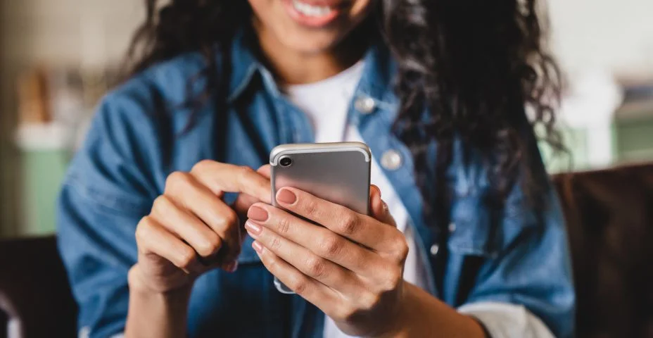 Foto cortada de uma jovem afro-americana usando telefone inteligente em casa. Mulher afro-americana sorridente usando smartphone em casa, mensagens ou navegando nas redes sociais enquanto relaxa no sofá