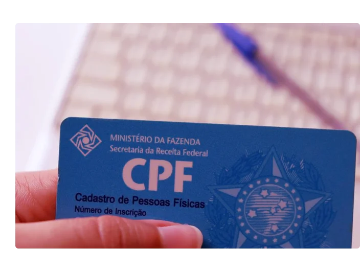 uma pessoa segurando cartão CPF no fundo tem um teclado de computador e uma caneta azul