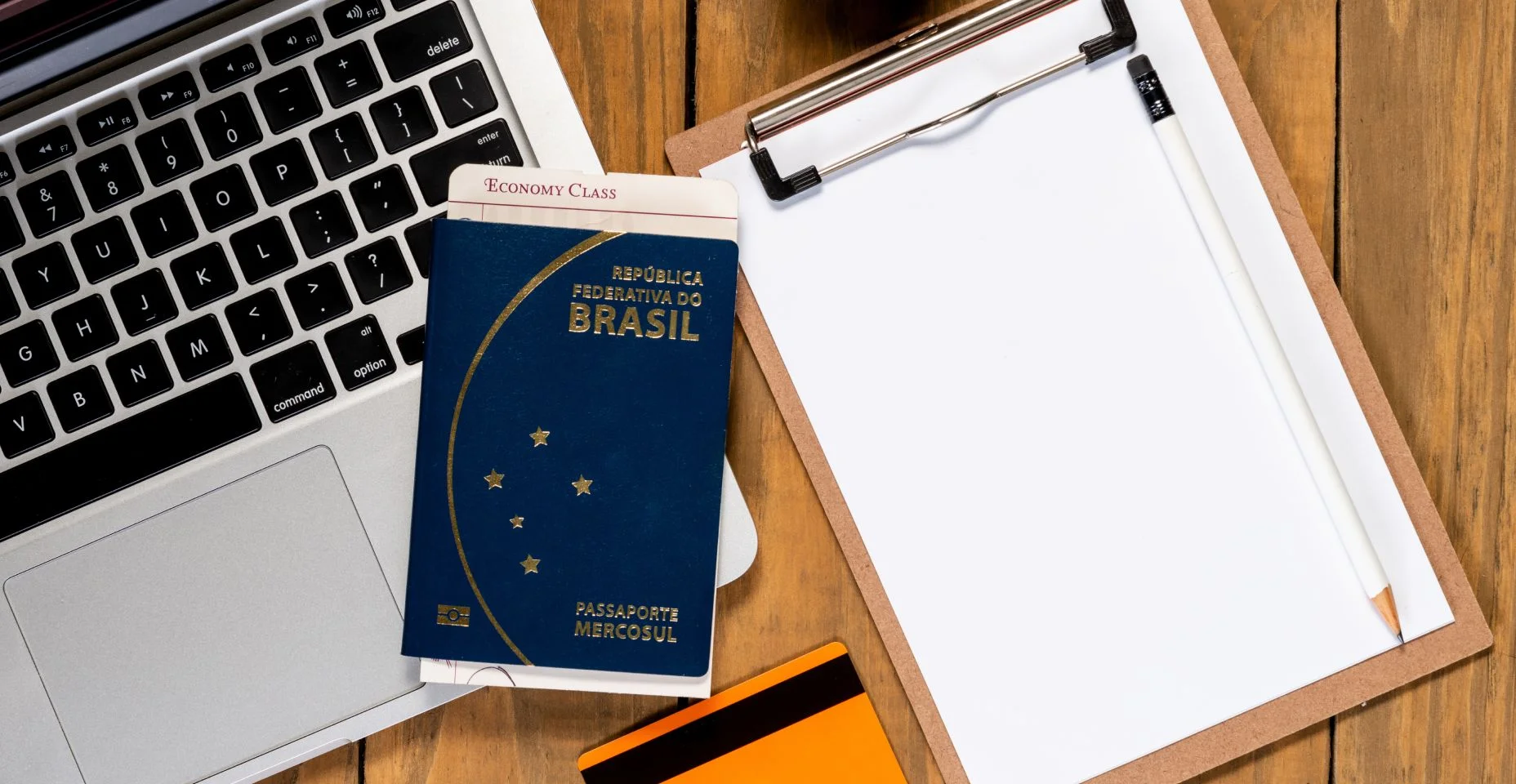 Passaporte brasileiro e passagem no computador na mesa de madeira. Bloco de notas em branco para planejamento de viagens.