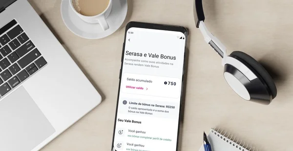 Um smartphone sobre uma mesa de madeira exibe a tela do aplicativo "Serasa e Vale Bonus", mostrando um saldo acumulado de 750 pontos e opções para utilizar o saldo. Ao redor do celular, há um par de fones de ouvido preto e branco, um notebook aberto, uma xícara de café sobre um pires e um bloco de notas com uma caneta.