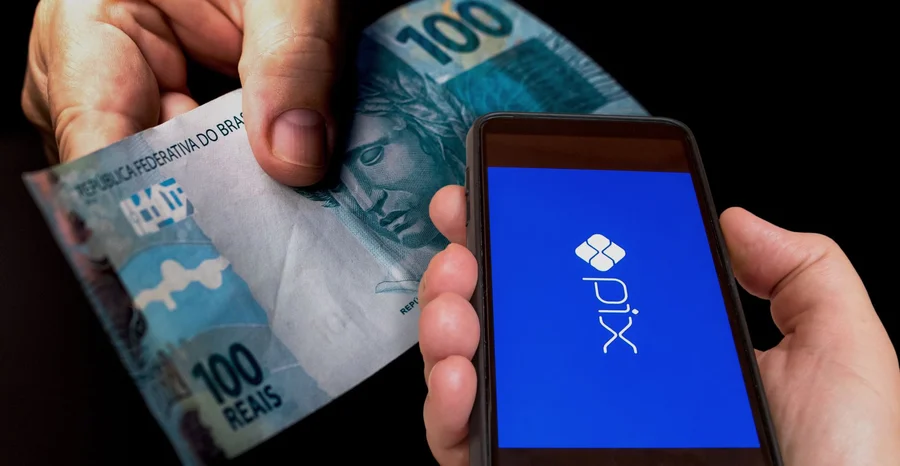 Tela do celular com sistema de pagamento instantâneo PIX do banco central do Brasil em fundo escuro. Close da mão com nota de cem reais.