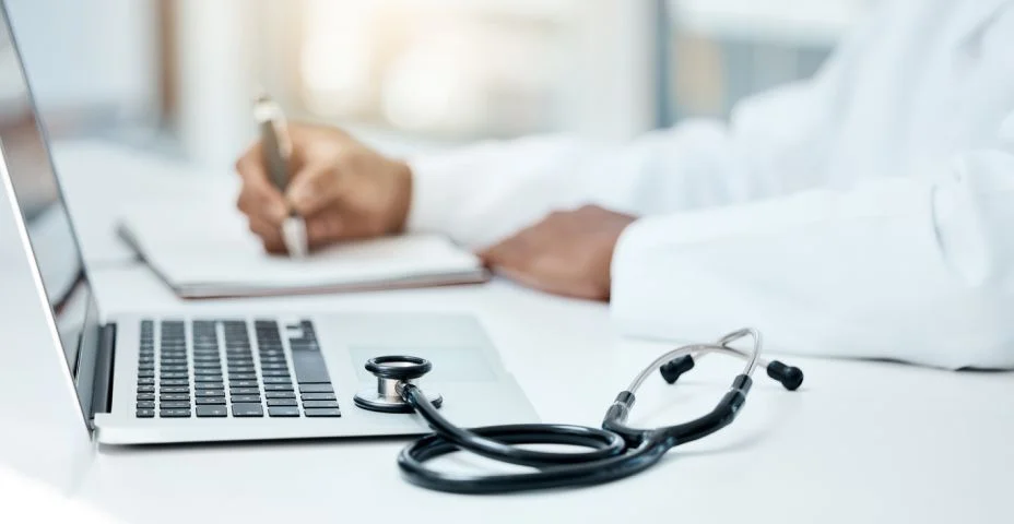 Uma mesa com notebook, estetoscópio e um médico assinando um papel