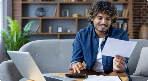 Homem novo que faz a papelada que senta-se na sala de visitas do apartamento no sofá, pagando cheques de crédito e pagamentos do aluguel, usando o portátil e a calculadora