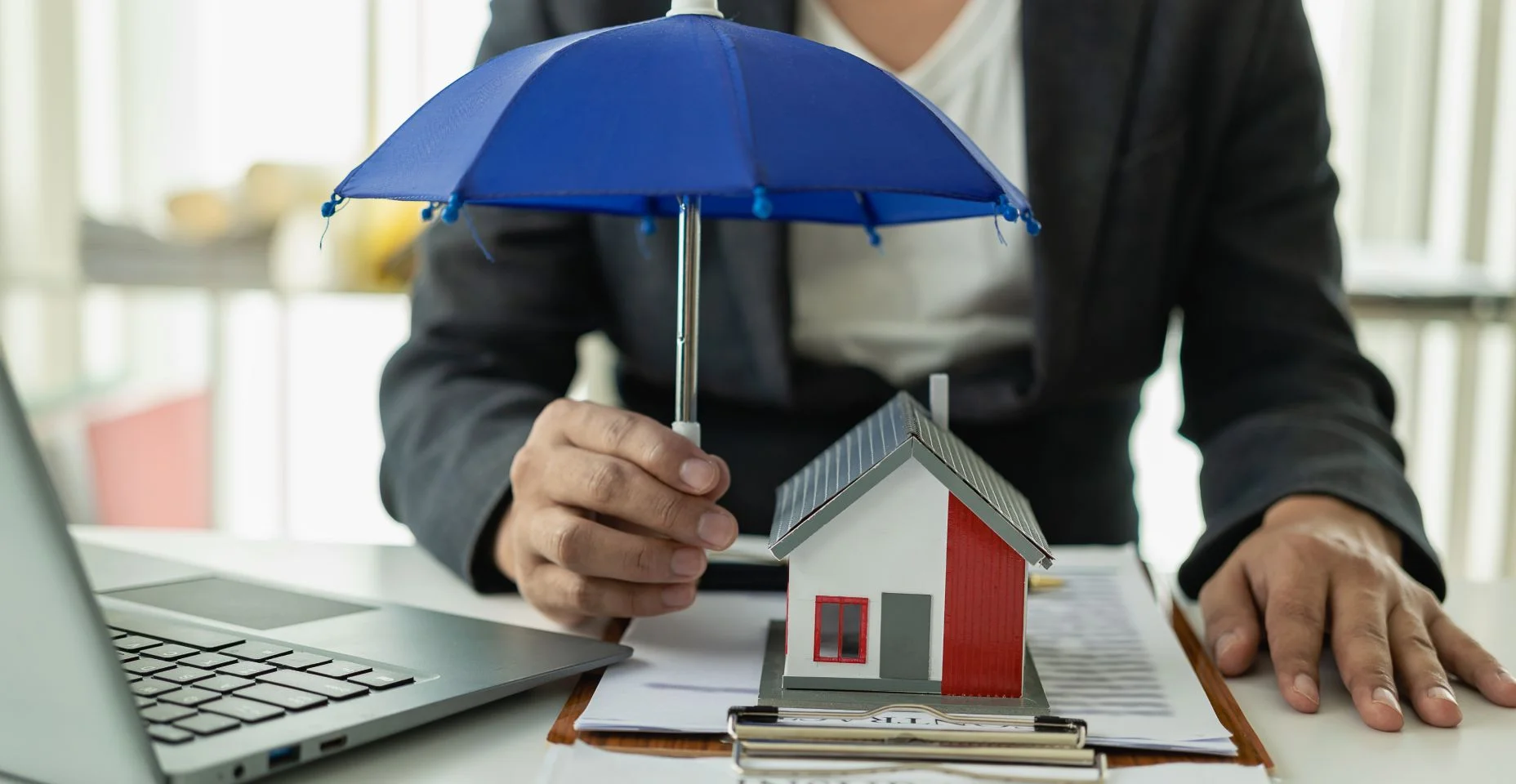 Guarda-chuva azul cobrindo um modelo de casa em uma mesa de madeira Seguro imobiliário, conceito de seguro hipotecário Seguro é o controle de risco no negócio imobiliário.