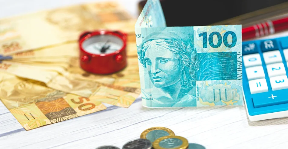 Notas e moedas de reais sobre uma mesa de madeira. Uma calculadora na composição. Economia e finanças brasileiras.