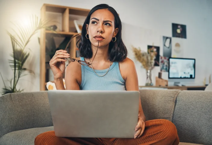 Mulher sentada no sofá com o notebook no seu colo e fazendo uma cara de dúvida