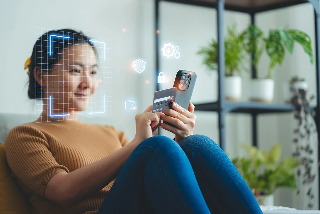 Mulher asiática segurando cartão de crédito e digitalização face id por telefone inteligente para compras on-line sentado no sofá na sala de estar em casa.