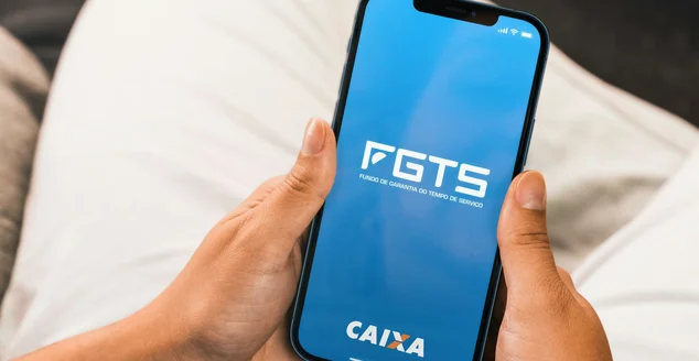 Mão feminina segurando um smartphone com aplicativo FGTS (Caixa) na tela.