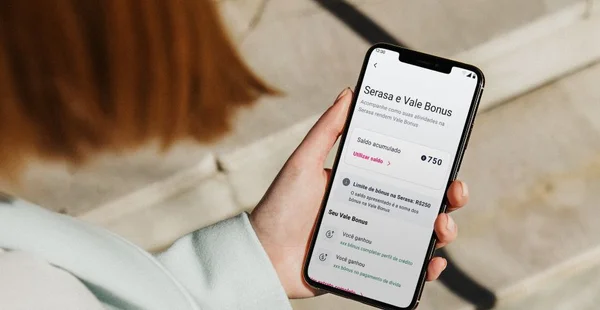 "Pessoa segurando um smartphone exibindo a tela de um aplicativo da Serasa com o título 'Serasa e Vale Bonus'. A tela mostra um saldo acumulado de 750 pontos, com opção de utilizar o saldo. Há informações sobre o limite de bônus de R$250 e detalhes dos bônus ganhos por completar o perfil de crédito e por pagamento em dia."