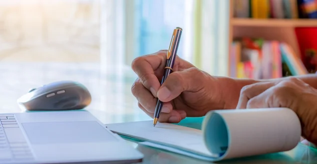 Mão de homem de negócios escrevendo e assinando talão de cheques com computador portátil na mesa de madeira no escritório doméstico moderno.