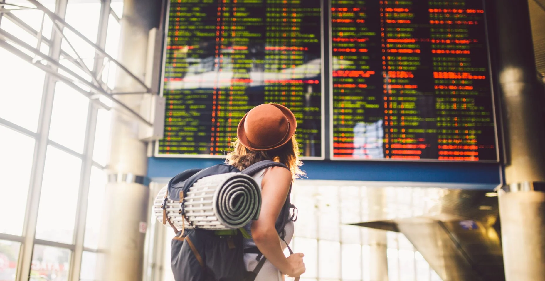 Tema viagens dos transportes públicos. jovem de pé com as costas no vestido e chapéu para trás a mochila e acampar equipamentos para dormir, esteira de isolamento parece horário na estação do Aeroporto de placar