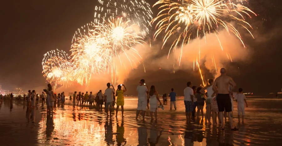 Celebração de Ano Novo na praia. Pessoas assistindo aos fogos de artifício à beira da água.