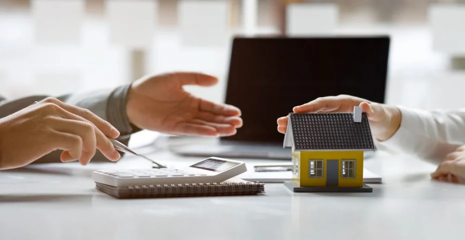 Duas mãos: uma segurando uma caneta e a outra gesticulando. Uma mesa com uma calculadora e um modelo em miniatura de uma casa. Ao fundo, um laptop aberto. Ambiente de escritório, possivelmente relacionado a finanças ou planejamento imobiliário.