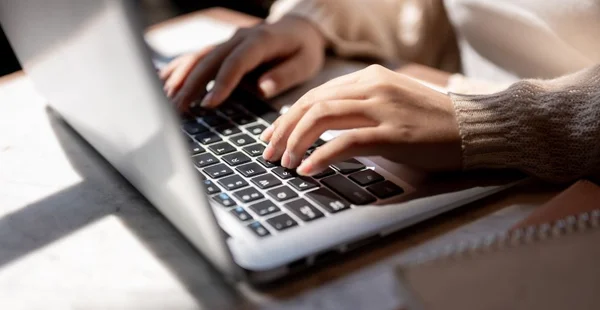 Imagem aproximada de uma mulher digitando no teclado, trabalhando em seu laptop em uma mesa dentro de casa.