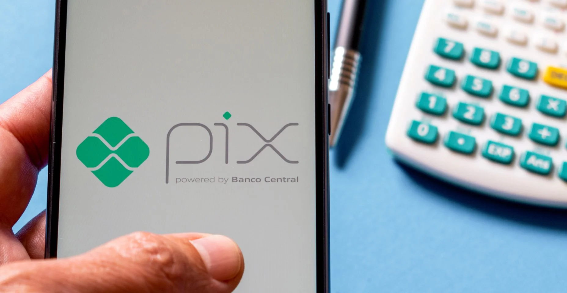 Mão segurando smartphone com aplicativo Pix, em cima da mesa com calculadora e caneta. Pix é o novo sistema de pagamentos instantâneos do Banco Central do Brasil.