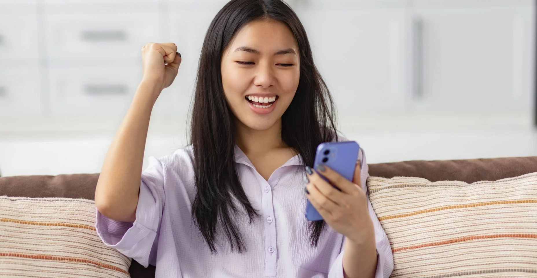 Animada jovem asiática feliz segurando o dispositivo de telefone inteligente, olhando para a tela do smartphone móvel gesticulando sim, ela recebeu boas notícias sentada no sofá em casa
