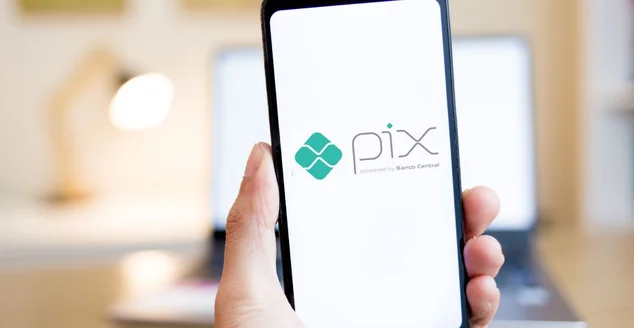 Mãos segurando um smartphone com o logotipo do PIX na tela com o interior de uma casa ao fundo. PIX é um sistema instantâneo de transferência e pagamento de dinheiro no Brasil.