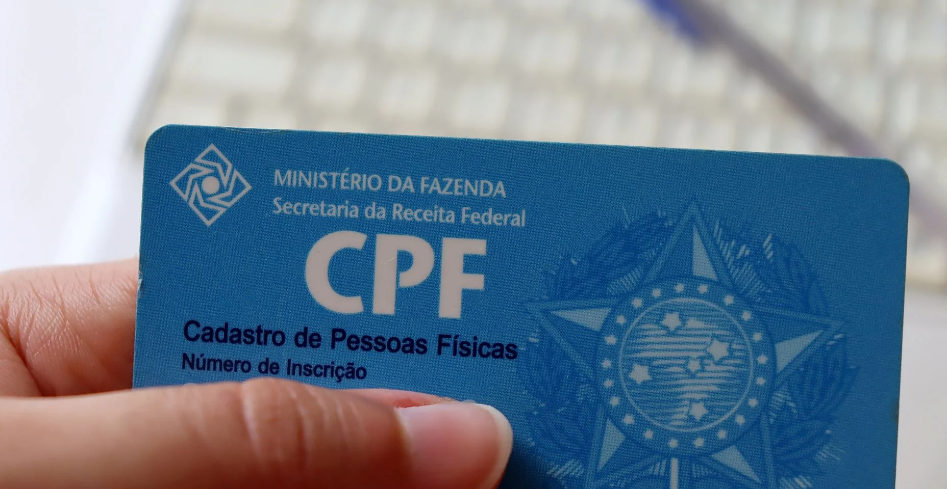 Pessoa segurando o CPF, com caneta e laptop ao fundo.